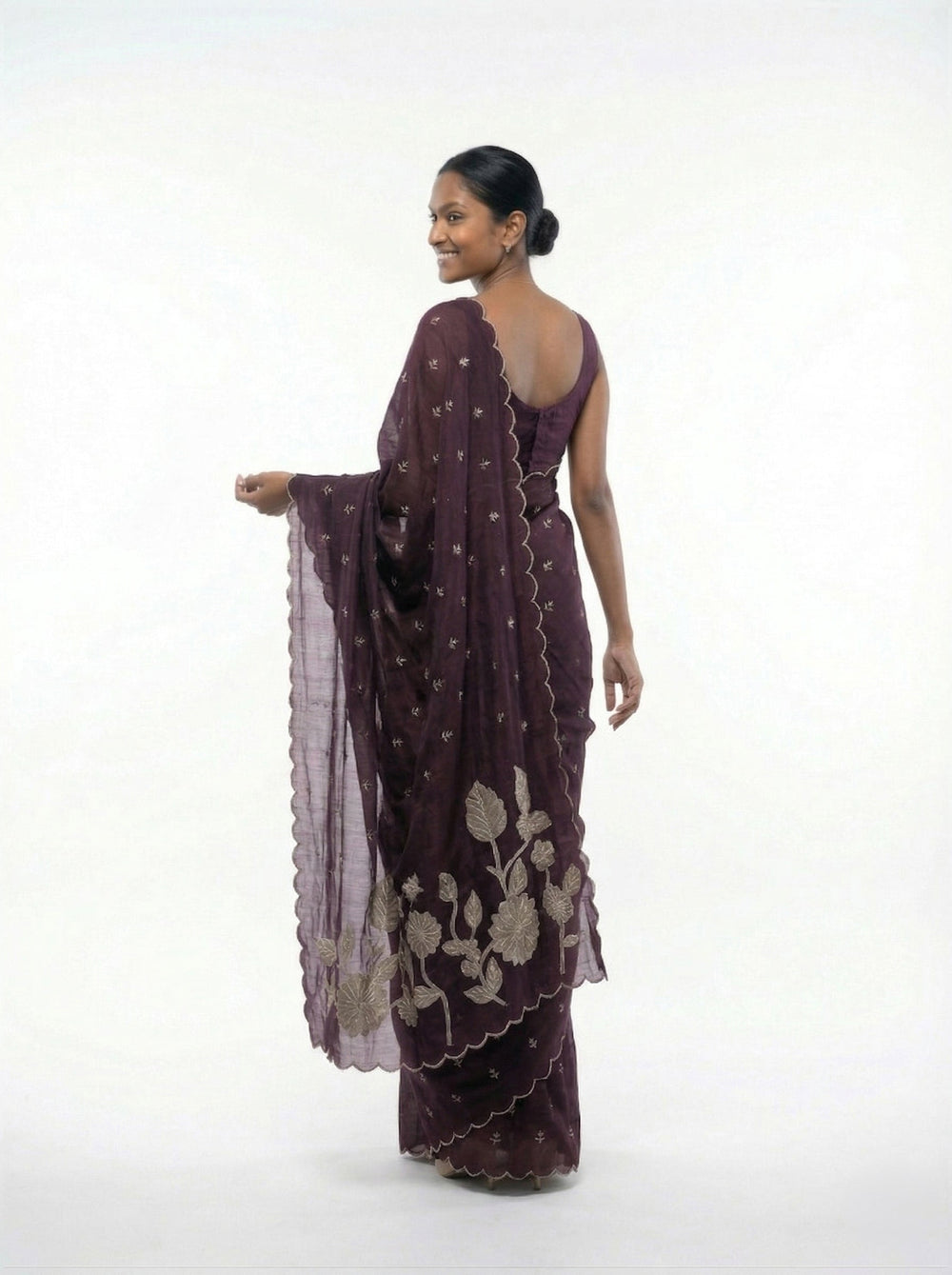 Woman in a purple saree standing in an outdoor setting with architectural elements and plants.