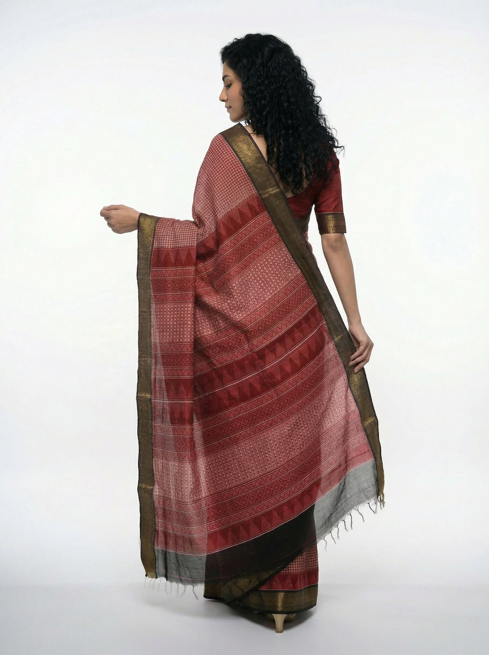 Woman in a red saree with gold border standing in front of a stone building.