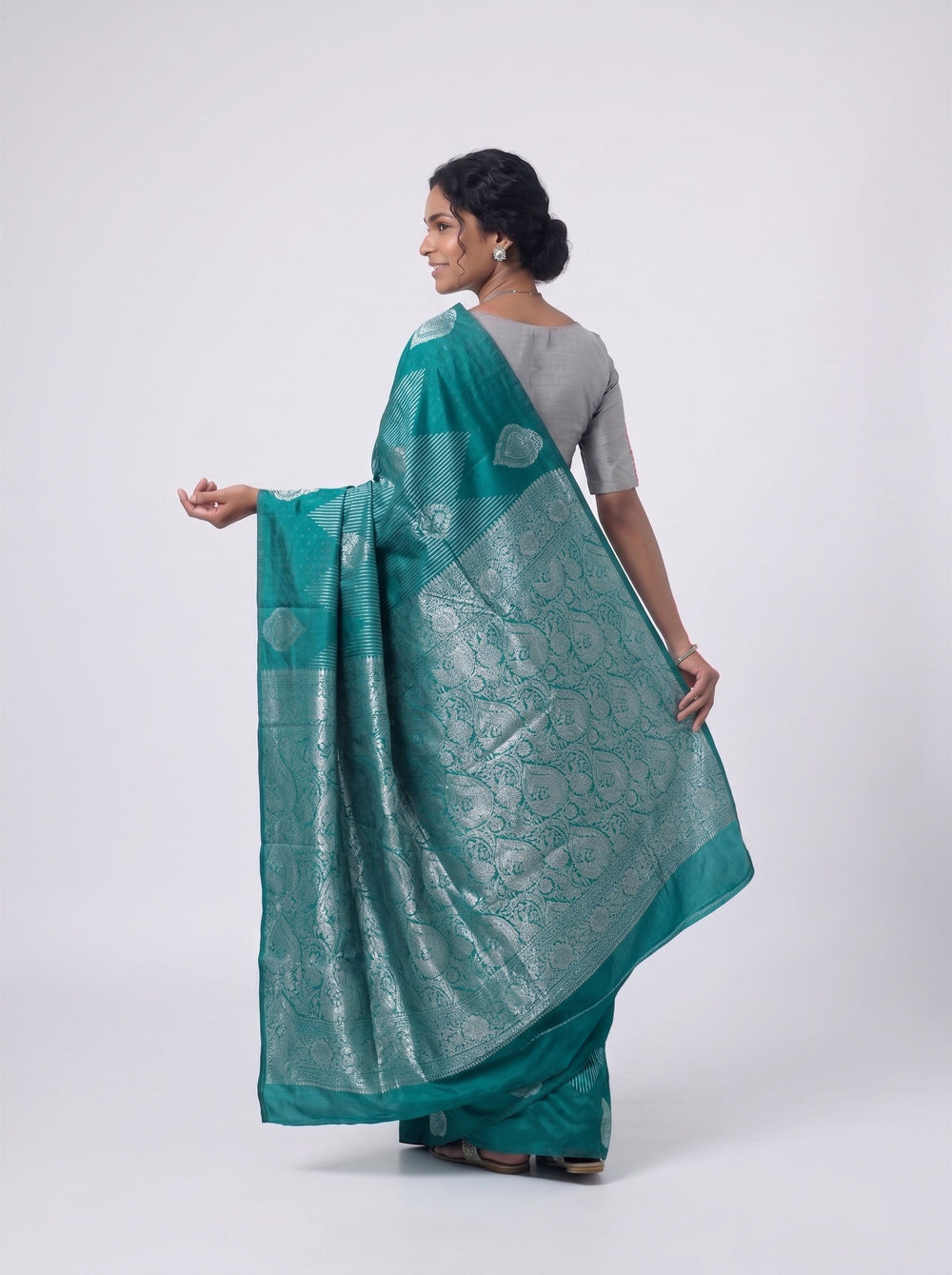 Woman in a teal saree standing in a room with a lamp and books.