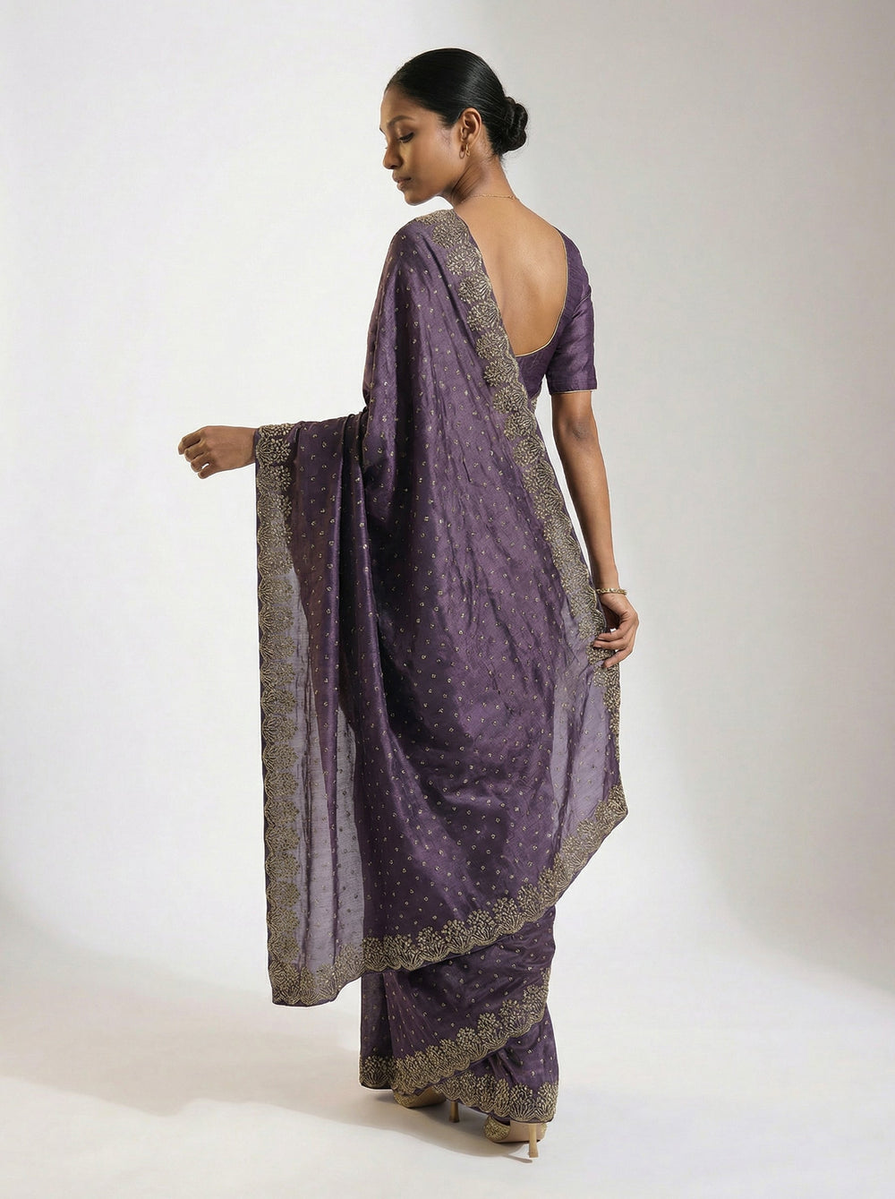 Woman in a purple saree standing in an outdoor setting with architectural elements and plants.