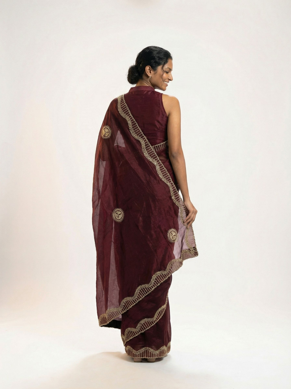 Woman in a maroon saree standing in an outdoor setting with plants and a building.
