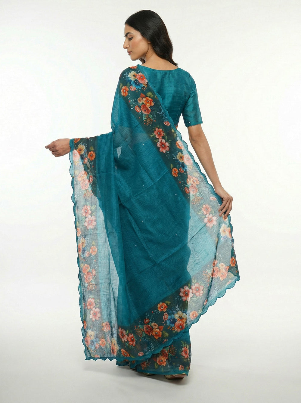 Woman in a teal saree with floral patterns standing in a room with a mirror and plants.