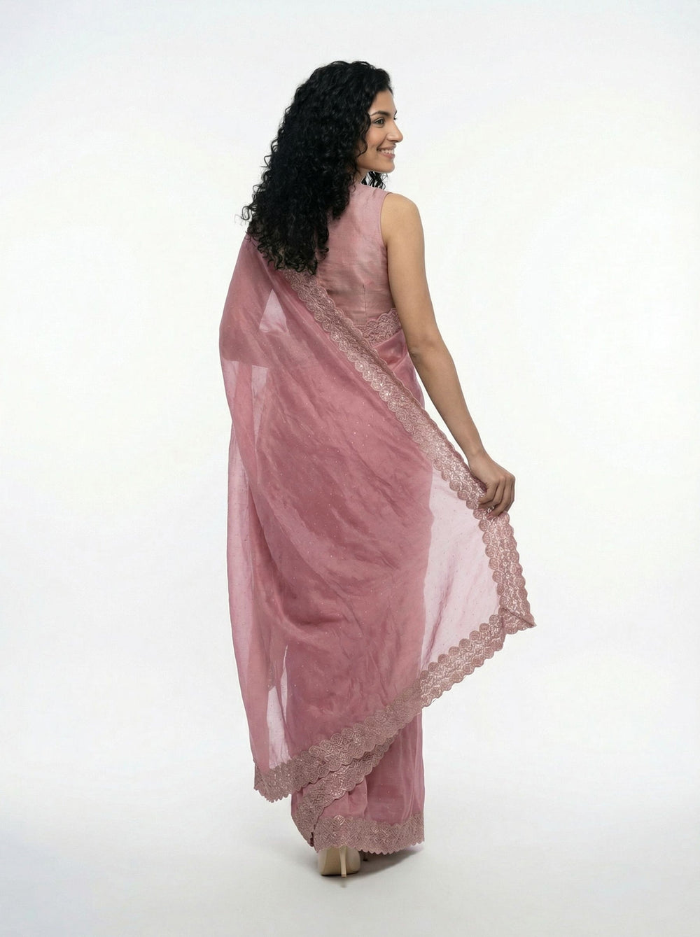 Woman in a pink saree standing in a garden setting with plants and stone architecture.