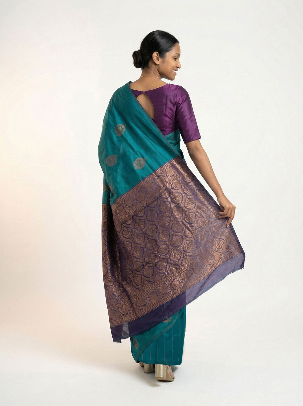 Woman in a teal saree with a purple blouse standing in a room with wooden furniture and books.