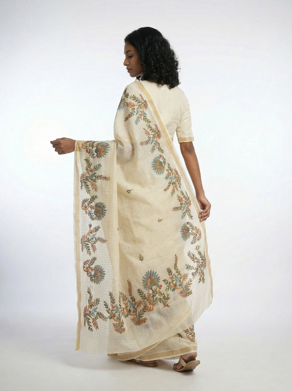 Woman in a cream saree with floral patterns standing in a traditional setting.