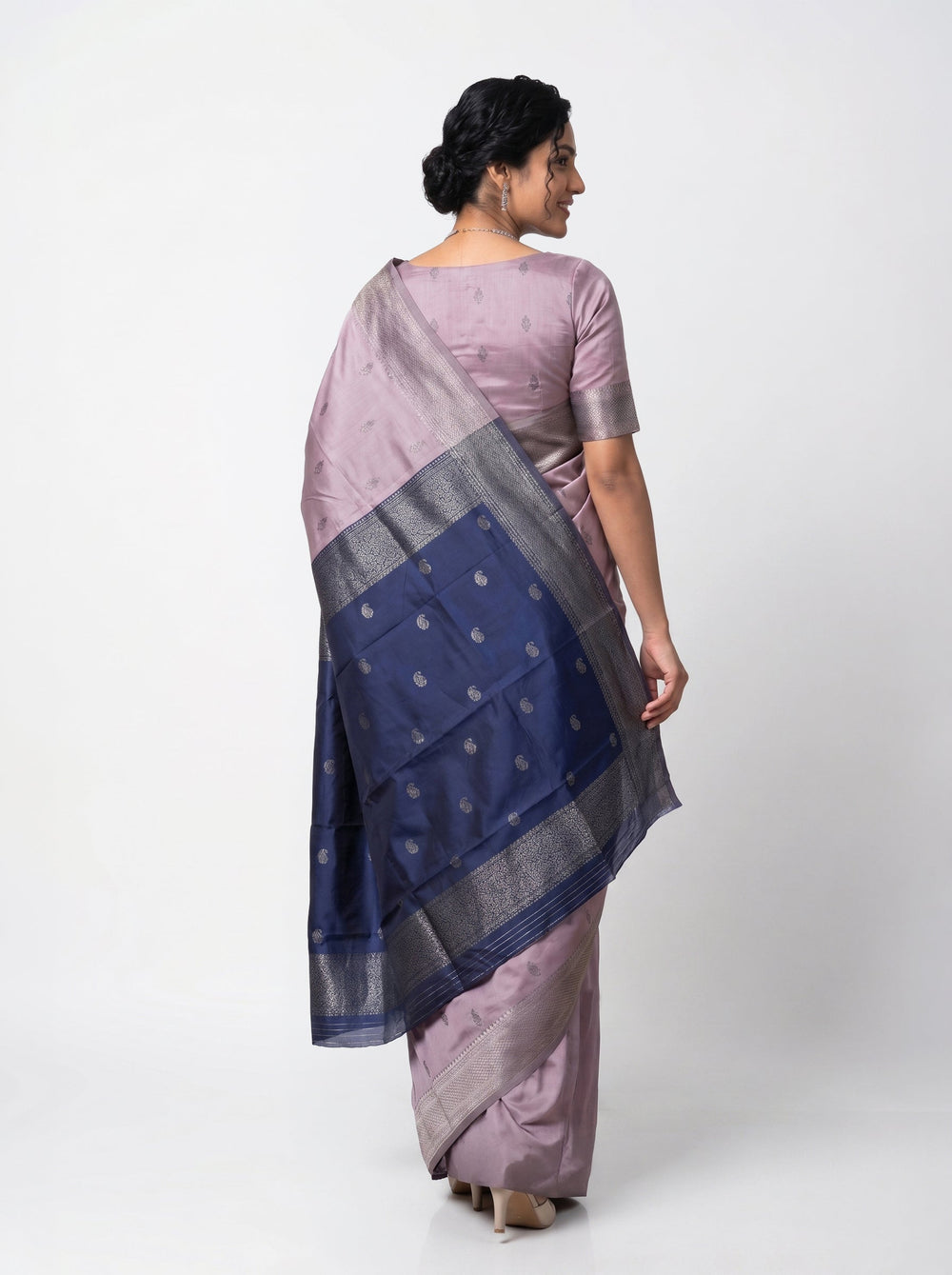 Woman in a lavender saree standing in a room with a desk and lamp.