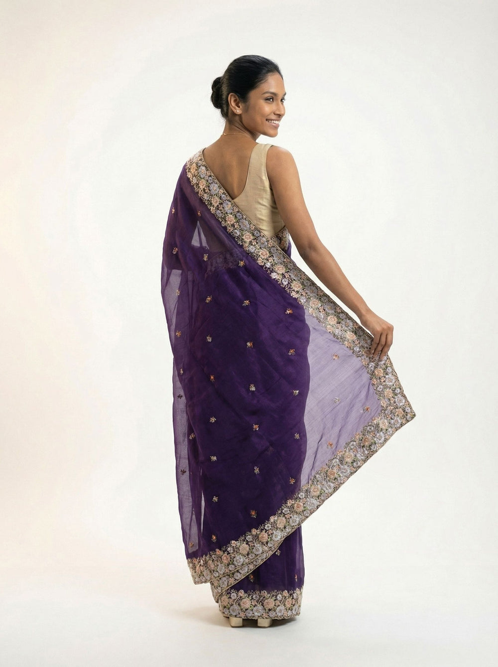 Woman in a purple saree standing in an outdoor setting with plants and architecture.
