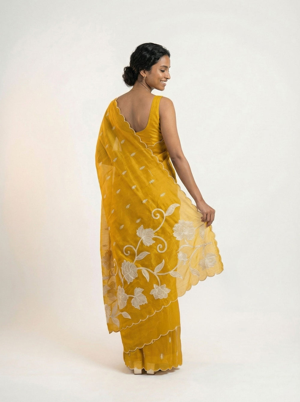Woman in a yellow saree standing in an outdoor setting with plants and architectural elements.