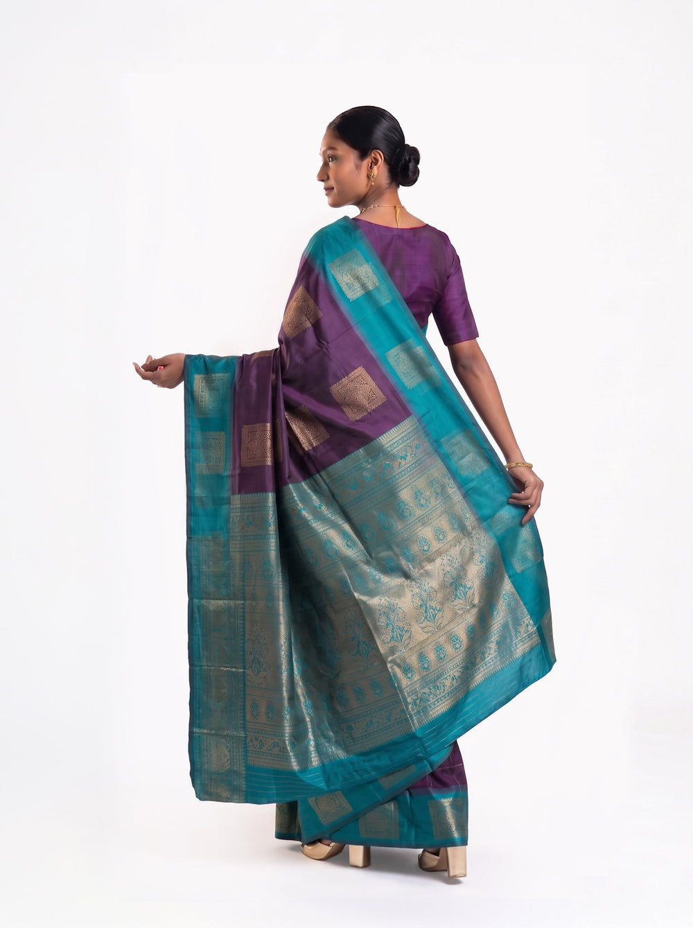 Woman in a purple and blue saree standing in a room with wooden furniture and decorative elements.