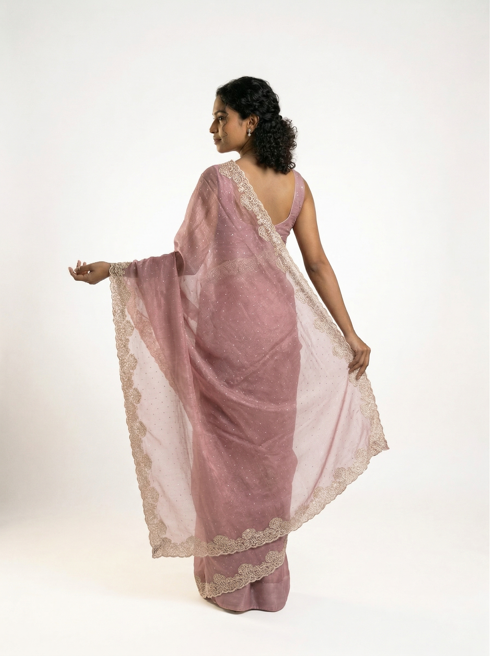 Woman in a pink saree standing outdoors near a traditional building with plants around.
