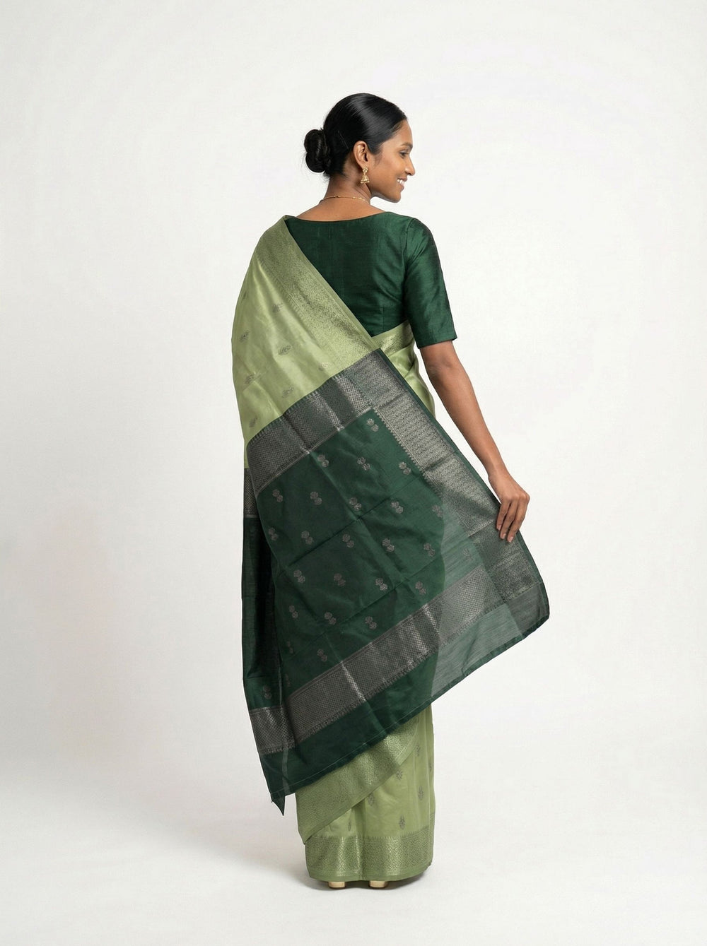 Woman in a green saree standing in a room with a bookshelf and lamp.