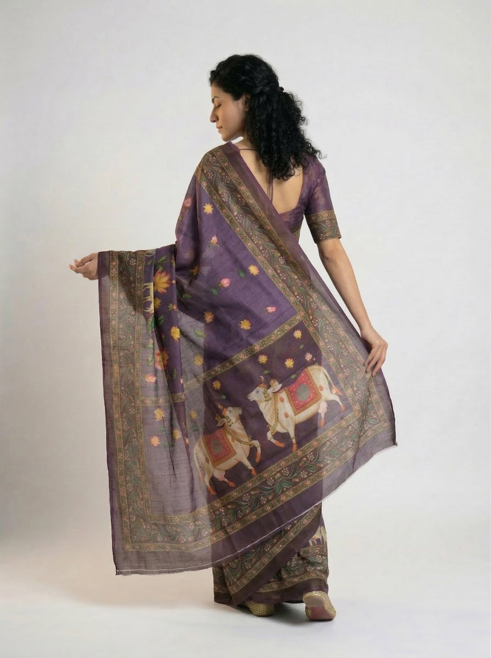 Woman in a traditional saree standing in a room with a lamp, plant, and framed picture.