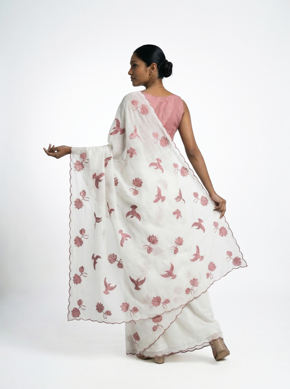 Woman in a white saree with pink floral patterns standing outdoors near a wooden pillar.