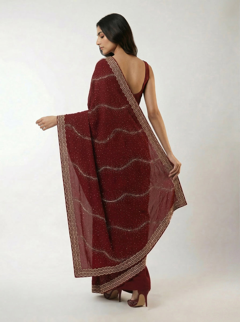 Woman in a maroon saree standing on a stone path with plants and a traditional building in the background.