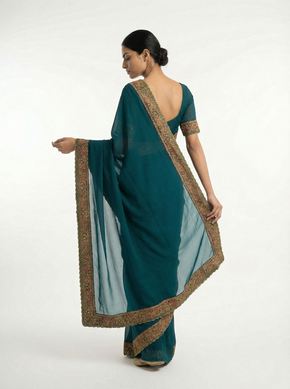 Woman in a teal saree standing on a balcony with architectural elements and plants in the background