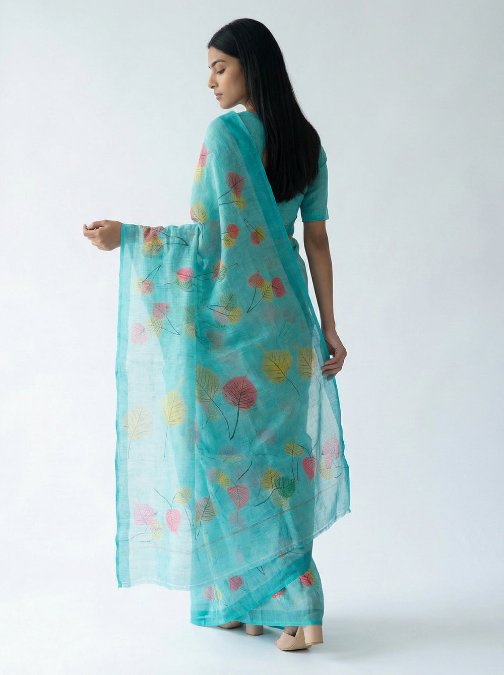 Woman in a turquoise saree with colorful patterns standing in a room with plants and a wooden cabinet.