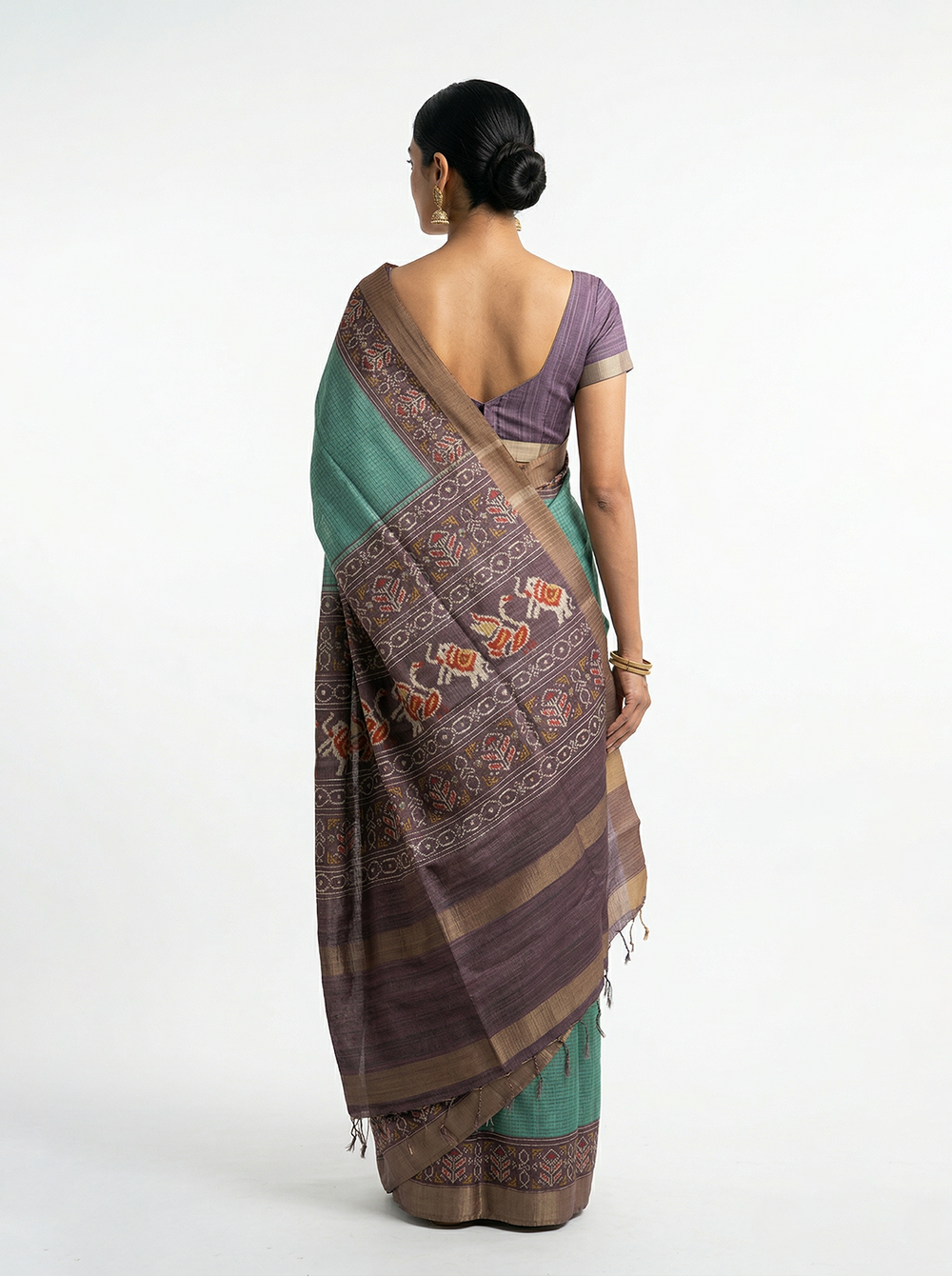 Woman in a green saree with a purple border standing in a hallway with plants.