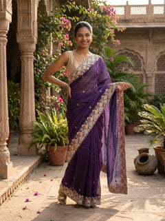 Woman in a purple saree standing in an outdoor setting with plants and architecture.