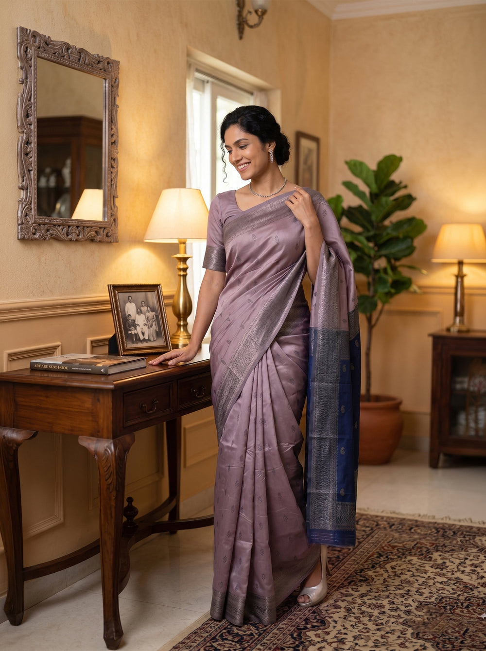 Woman in a lavender saree standing in a room with a desk and lamp.