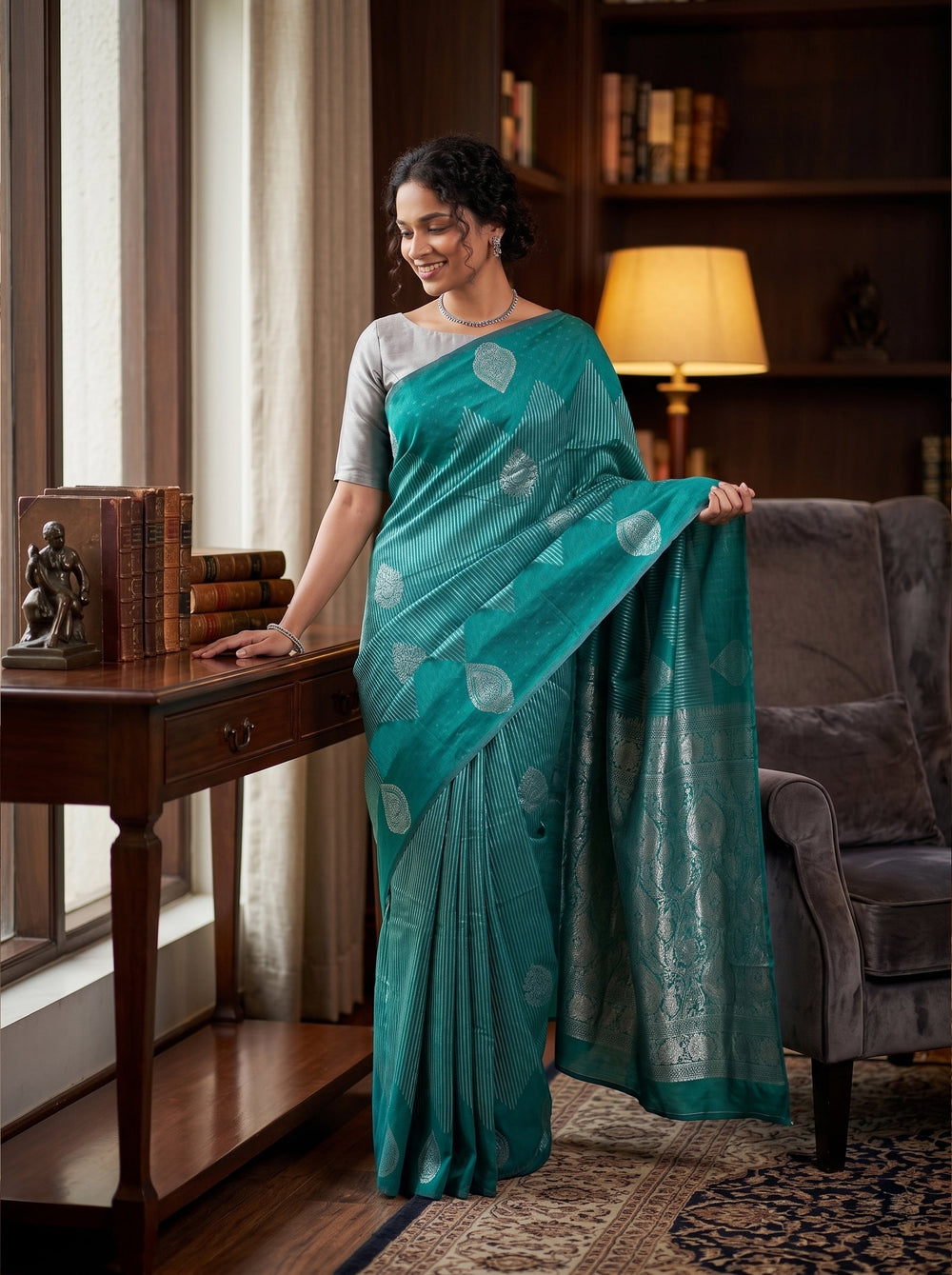 Woman in a teal saree standing in a room with a lamp and books.