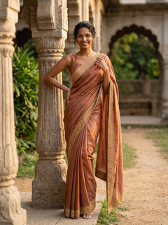 Woman in a traditional saree standing in an outdoor setting with stone architecture and greenery.