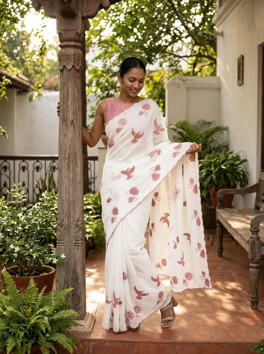 Woman in a white saree with pink floral patterns standing outdoors near a wooden pillar.