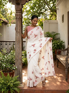 Woman in a white saree with pink floral patterns standing outdoors near a wooden pillar.