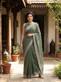 Woman in a green saree standing in an outdoor setting with plants and architectural elements.