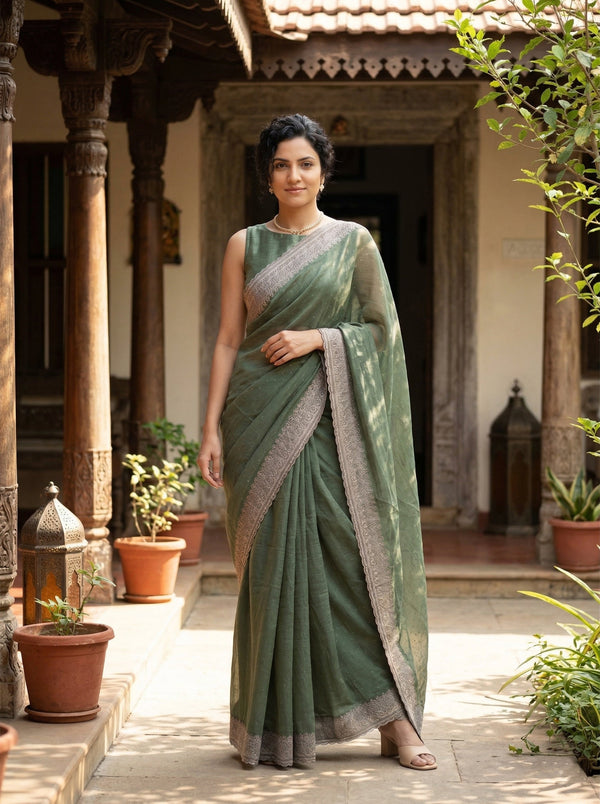 Woman in a green saree standing in an outdoor setting with plants and architectural elements.