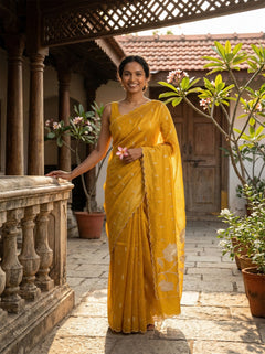Woman in a yellow saree standing in an outdoor setting with plants and architectural elements.