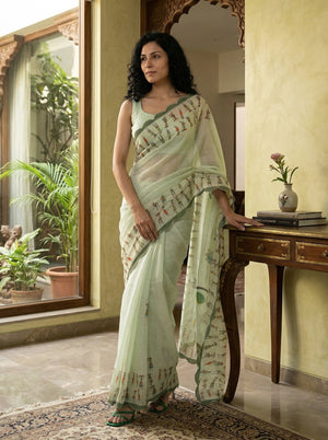 Woman in a light green saree standing in a room with a mirror and plants.