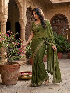 Woman in a green saree standing in an outdoor setting with plants and architectural elements.