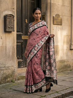 Woman in a pink saree with floral patterns standing in front of a building entrance.