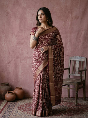 Woman in a patterned saree standing against a pink wall with rustic decor.