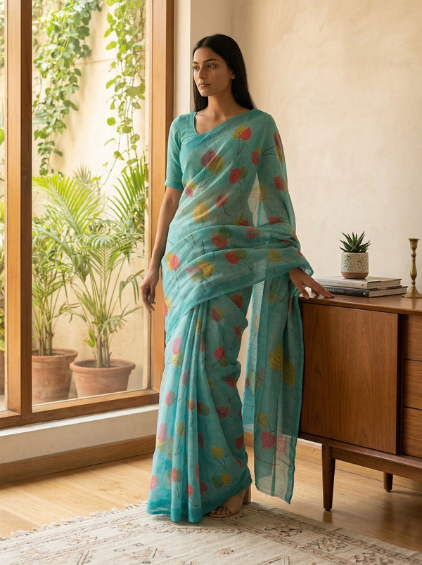 Woman in a turquoise saree with colorful patterns standing in a room with plants and a wooden cabinet.