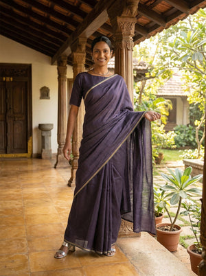 Woman in a dark blue saree standing in a traditional setting with plants and wooden architecture.