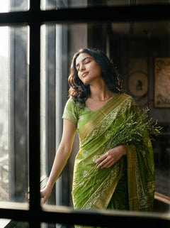 Woman in a green saree holding flowers, standing indoors with large windows.