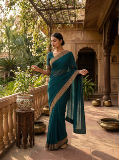 Woman in a teal saree standing on a balcony with architectural elements and plants in the background