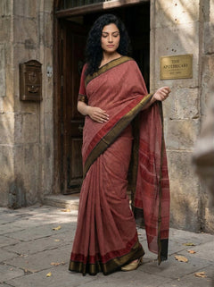 Woman in a red saree with gold border standing in front of a stone building.