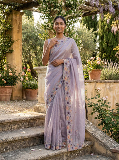 Woman in a lavender saree with floral patterns standing in an outdoor garden setting.