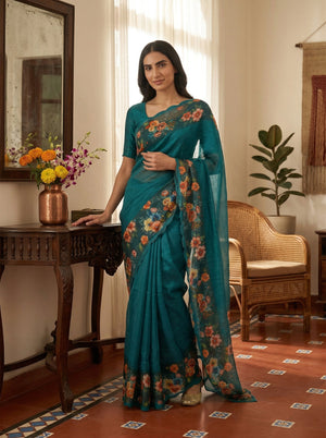Woman in a teal saree with floral patterns standing in a room with a mirror and plants.
