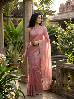 Woman in a pink saree standing in a garden setting with plants and stone architecture.