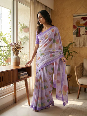Woman in a light purple saree with floral patterns standing in a bright room with large windows.