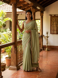 Woman in a light green saree standing in a traditional setting with wooden pillars and plants.