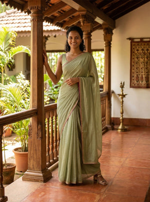 Woman in a light green saree standing in a traditional setting with wooden pillars and plants.