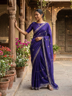 Woman in a purple saree standing in an outdoor setting with flowers and architectural elements.