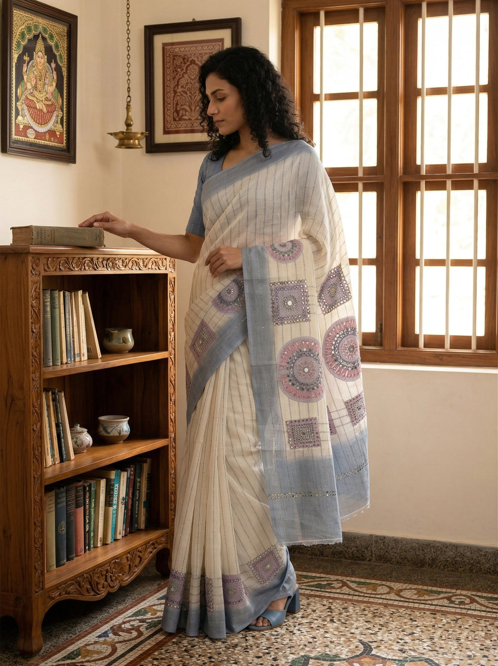 Woman in a saree standing in a room with wooden bookshelf and window