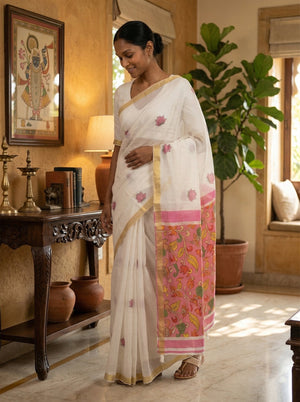 Woman in a white saree with pink border standing in a room with decor elements.