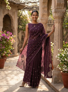 Woman in a purple saree standing in an outdoor setting with architectural elements and plants.