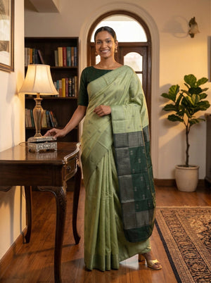 Woman in a green saree standing in a room with a bookshelf and lamp.